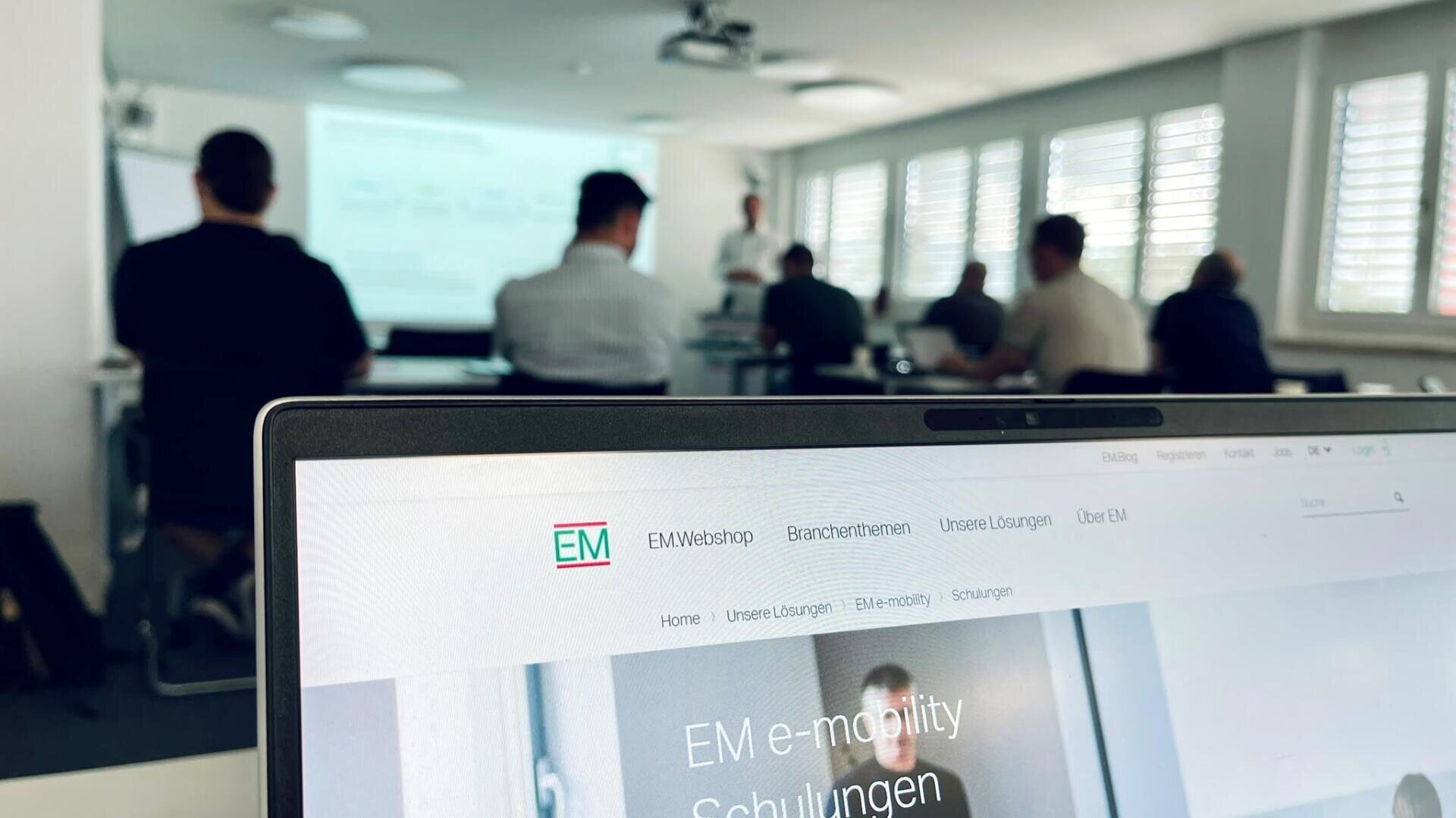 Im Vordergrund steht ein Laptop, auf dem eine Website zur E-Mobilitätsschulung angezeigt wird, während im Hintergrund mehrere Personen in einem hellen, modernen Unterrichtsraum einer Präsentation beiwohnen.