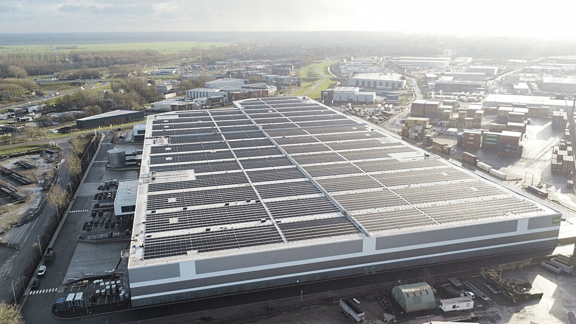 Ein großes Industrielager mit einem mit Sonnenkollektoren bedeckten Dach, umgeben von Parkplätzen, Containern und anderen Industriegebäuden unter einem bewölkten Himmel.