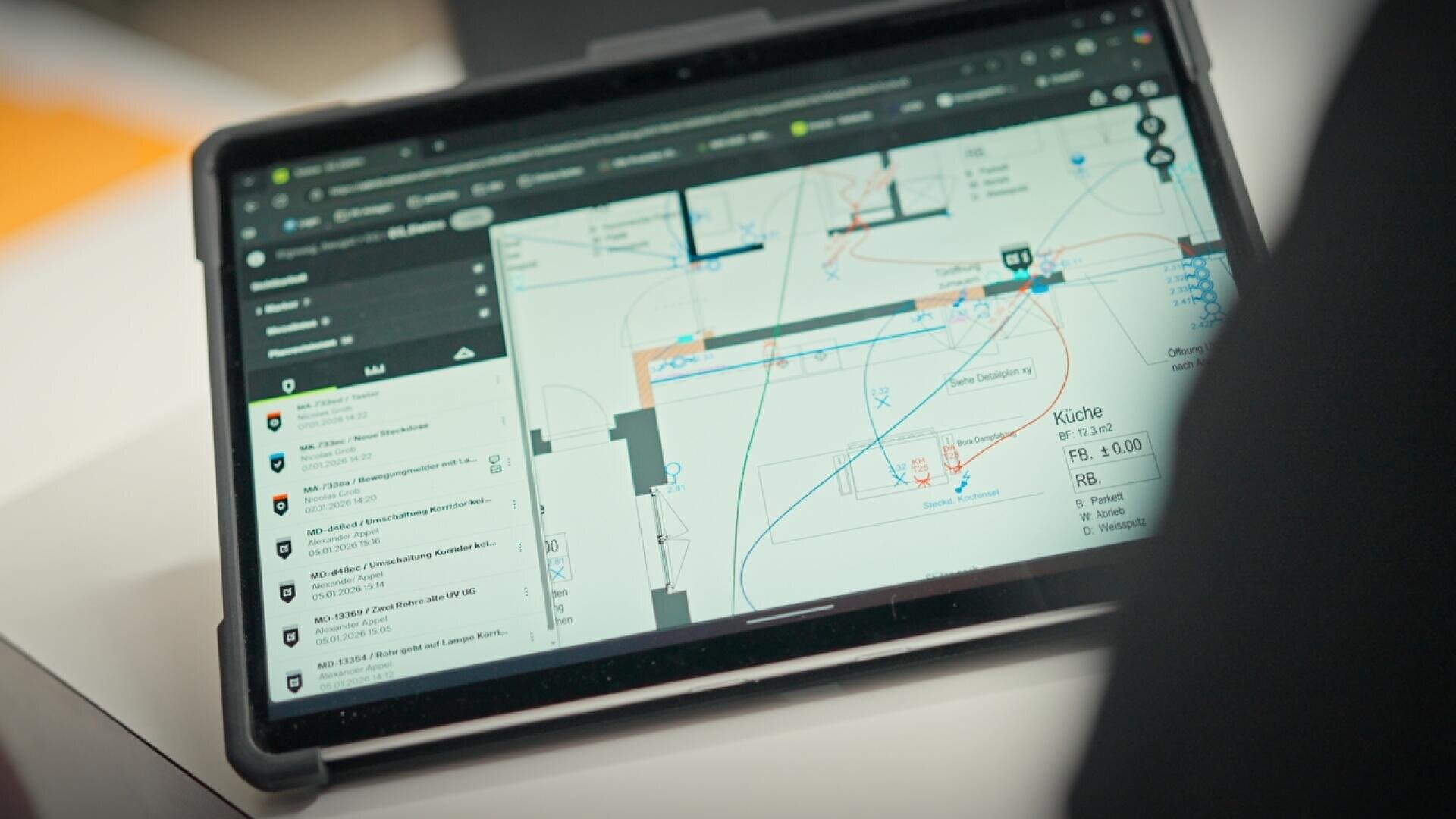 Ein Tablet zeigt eine technische Zeichnung oder einen Bauplan mit verschiedenen farbigen Linien und Symbolen sowie eine Seitenleiste mit einer Liste von Dokumenten oder Benachrichtigungen. Das Gerät ruht auf einer weißen Oberfläche.