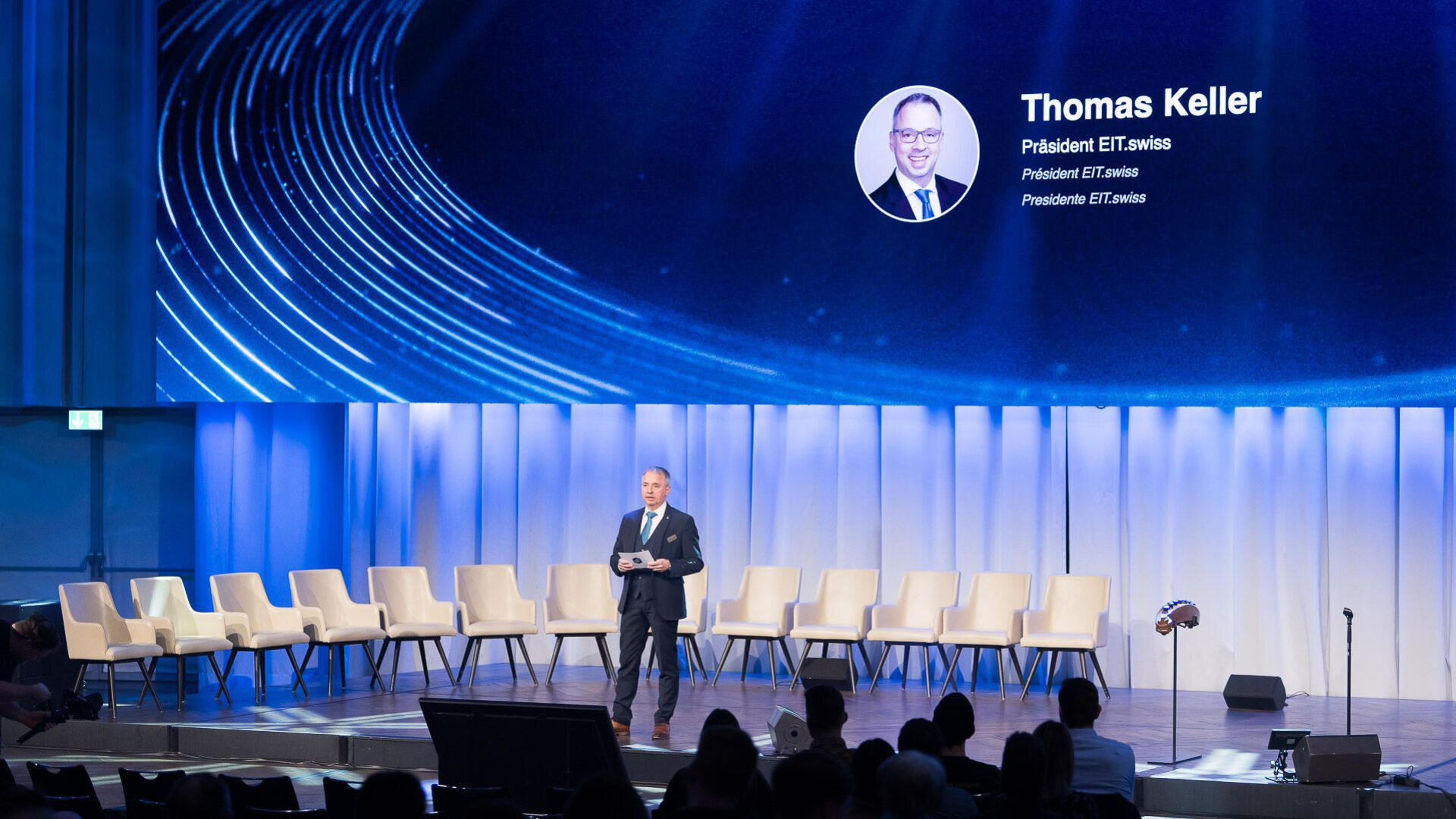 Ein Mann steht auf der Bühne und hält Papiere vor leeren Stühlen in der Hand. Hinter ihm ist ein großer Bildschirm mit einem Foto und dem Namen Thomas Keller, Président EIT.swiss, in einem modernen Auditorium zu sehen.