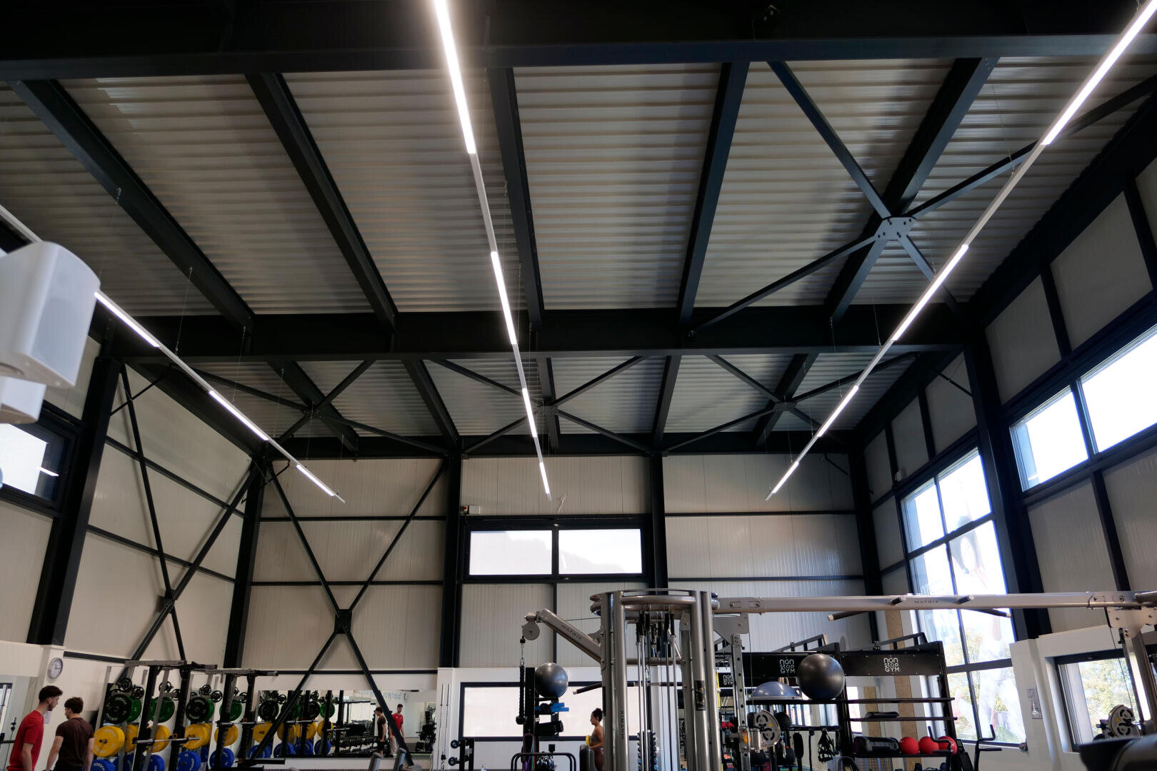 Ein modernes Fitnessstudio mit hohen Decken, freiliegenden Balken, großen Fenstern, Hantelständern, Fitnessgeräten und ein paar Trainierenden. Helle LED-Lichter laufen über die Decke.