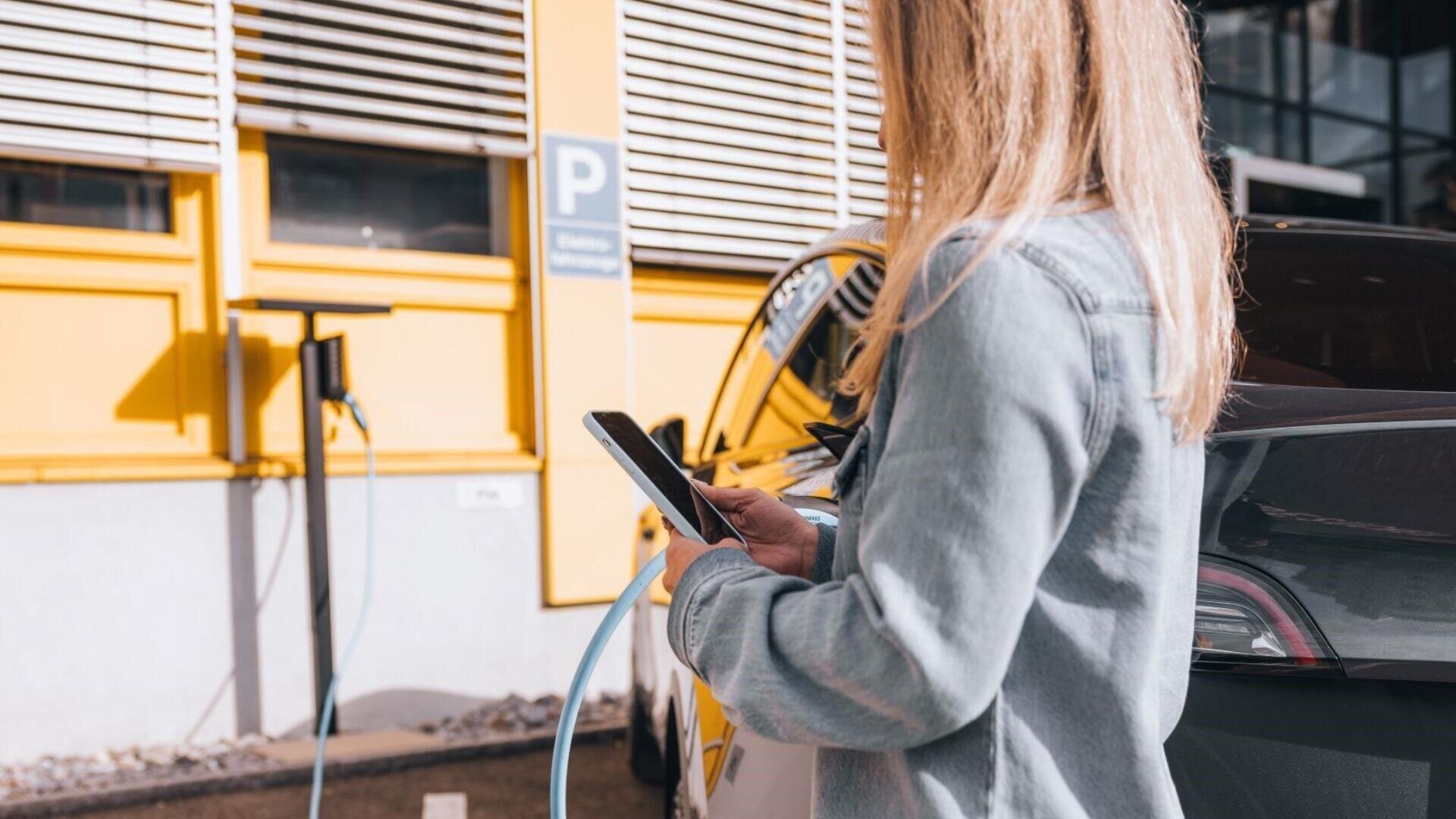 Eine Person mit langen Haaren benutzt ein Smartphone, während sie neben einem schwarzen Elektroauto steht, das an einer Ladestation vor einem gelben Gebäude angeschlossen ist.