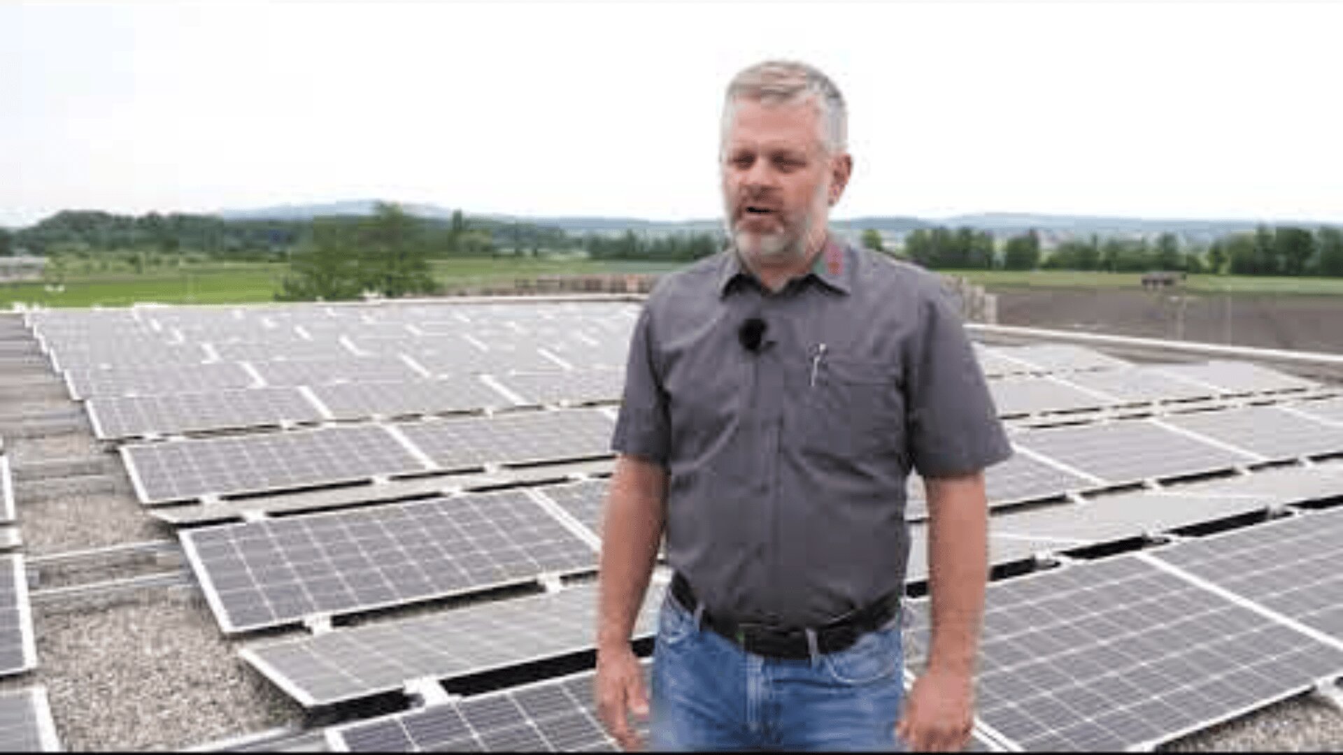 Ein Mann mit grauen Haaren und Bart steht vor Reihen von Solarmodulen auf einem Dach, mit einer grünen Landschaft und einem bewölkten Himmel im Hintergrund.