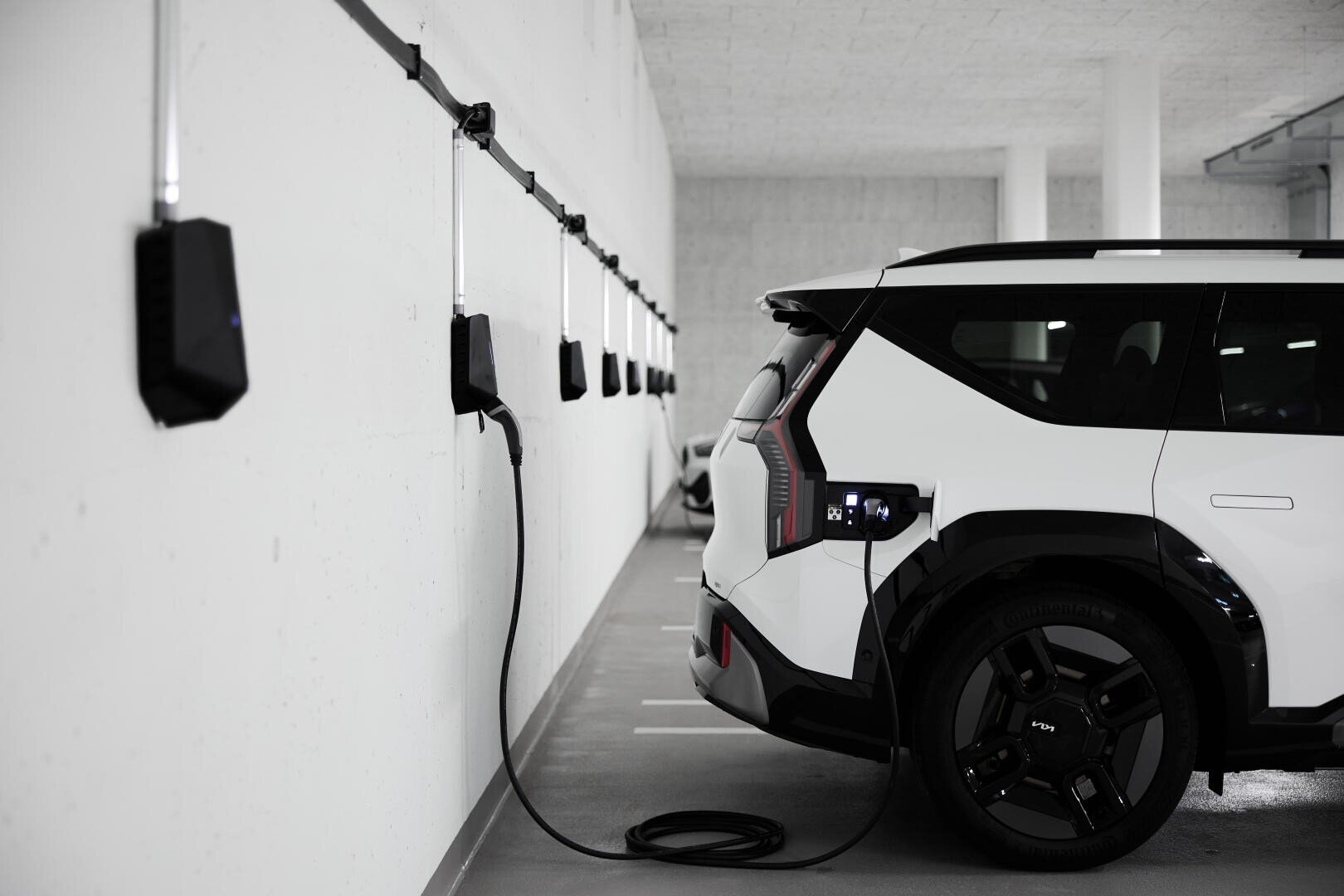 Un SUV électrique blanc est garé dans un garage et branché à une borne de recharge murale. On aperçoit plusieurs autres bornes de recharge le long du mur.