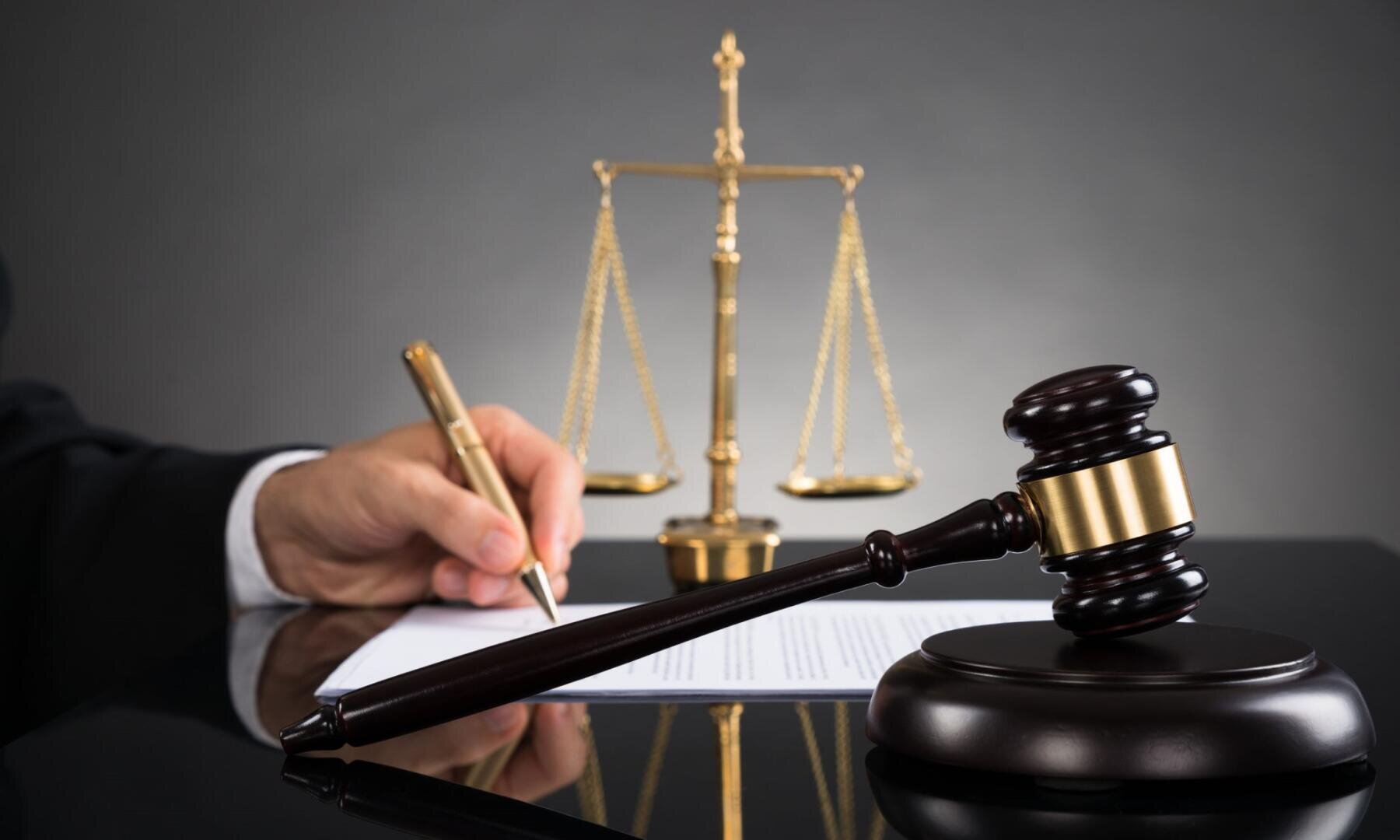 Eine Person im Anzug unterschreibt an einem Schreibtisch mit einem goldenen Stift ein Dokument. Im Vordergrund sind ein Richterhammer aus Holz und eine Waage der Gerechtigkeit aus Messing zu sehen, die für Recht und Gerechtigkeit stehen.