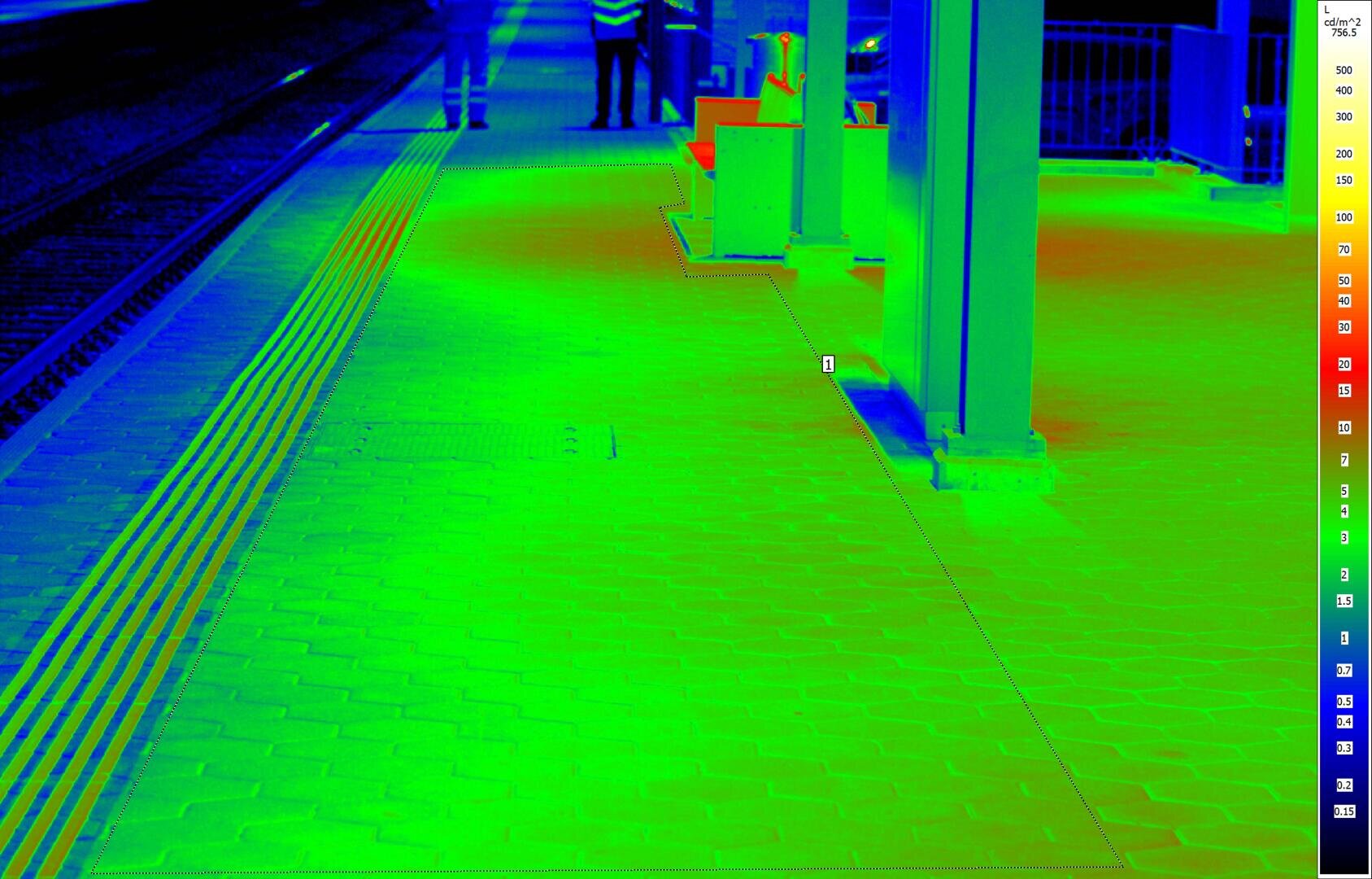Wärmebild eines Bahnsteigs, das Temperaturunterschiede von blau (kälter) bis rot (wärmer) zeigt, mit zwei Personen und den Bahnhofsgebäuden im Hintergrund; rechts ist eine Temperaturskala zu sehen.