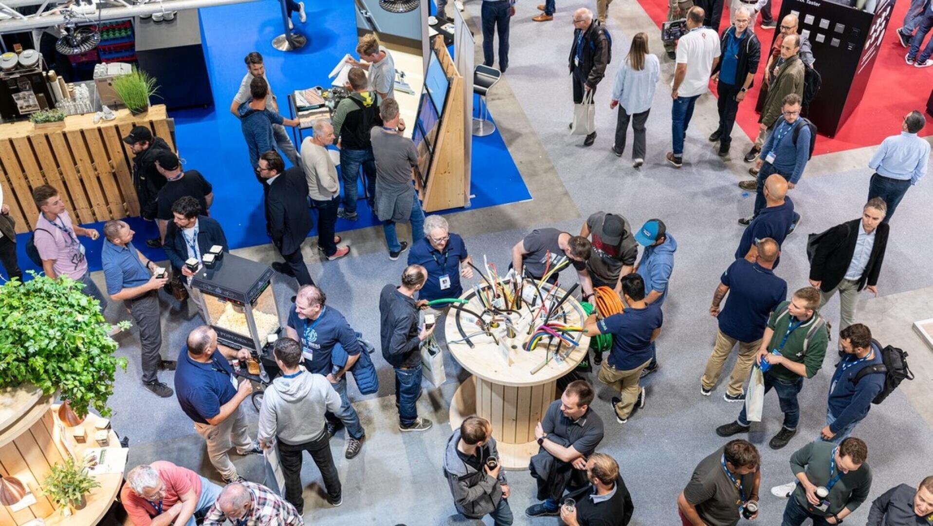 Auf einer Messe versammelt sich eine Menschenmenge um Ausstellungsstände und Tische mit Pflanzen und bunten Kabeln und tauscht sich in einem großen Innenraum aus und knüpft Kontakte.