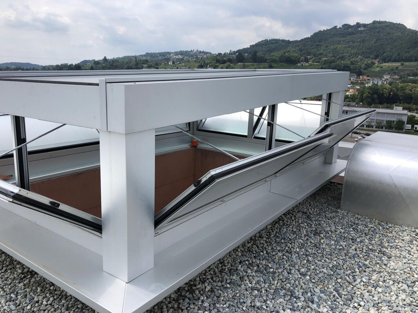 Auf einem mit Kies bedeckten Dach ist eine rechteckige Dachfensterkonstruktion aus Metall mit offenen Glasflächen angebracht. Im Hintergrund sind unter einem bewölkten Himmel Hügel und Gebäude zu sehen.