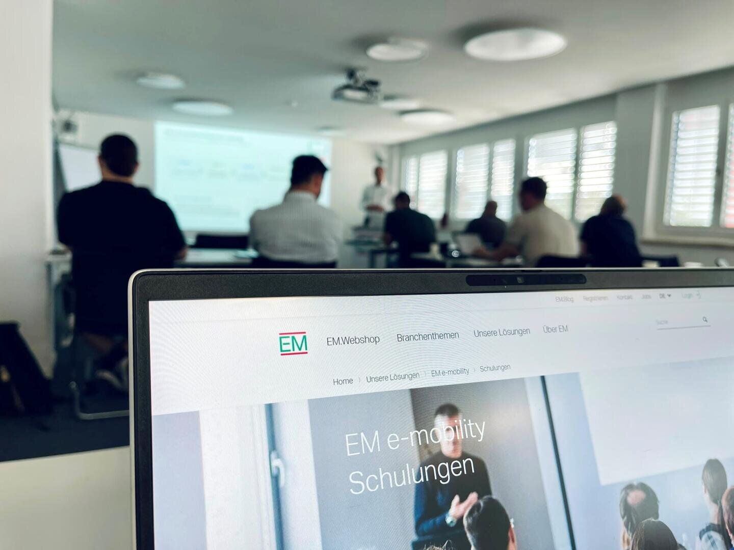 Au premier plan, un ordinateur portable affiche le site web de formation à la mobilité électrique « EM », tandis qu'à l'arrière-plan, plusieurs personnes assistent à une présentation dans une salle de classe bien éclairée.
