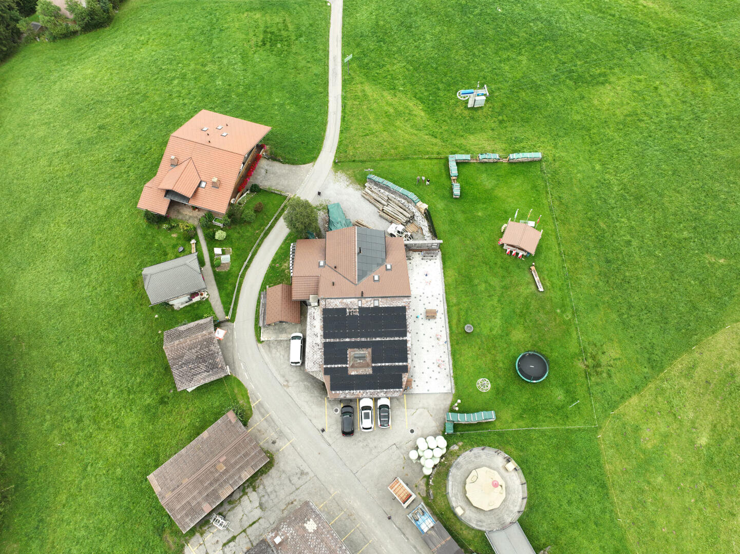 Luftaufnahme eines ländlichen Grundstücks mit mehreren Gebäuden, gepflasterten Straßen, grünem Gras, einem kreisförmigen Bereich, einem Trampolin, einer Spielplatzrutsche und geparkten Fahrzeugen. Das Gelände ist von offenen Feldern umgeben.