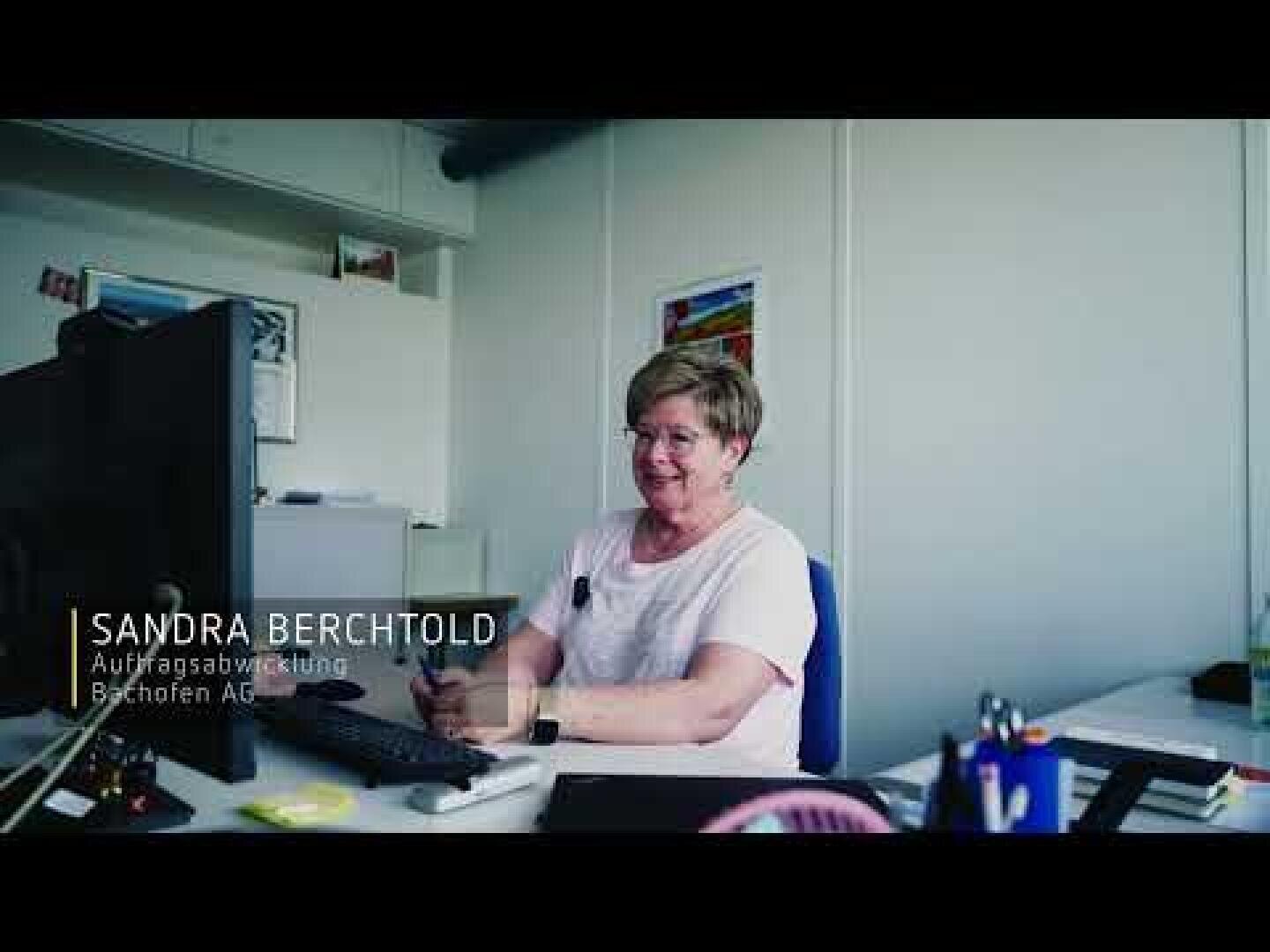 Eine Frau mit kurzen Haaren und Brille, die ein hellrosa Oberteil trägt, sitzt an ihrem Schreibtisch in einem hellen Büro und lächelt. Auf dem Bild steht: Sandra Berchtold, Auftragsabwicklung, Buchsifen AG.