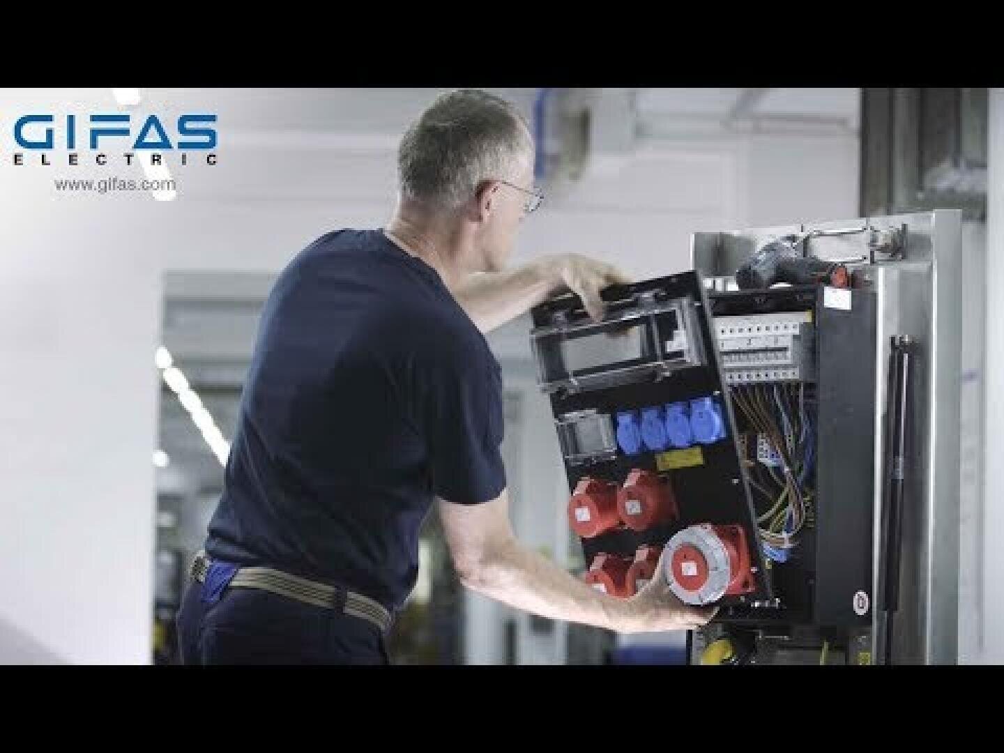 Un homme portant des lunettes travaille sur un tableau électrique ouvert avec plusieurs prises et câbles de couleur. Le logo de GIFAS Electric est visible dans le coin supérieur gauche.