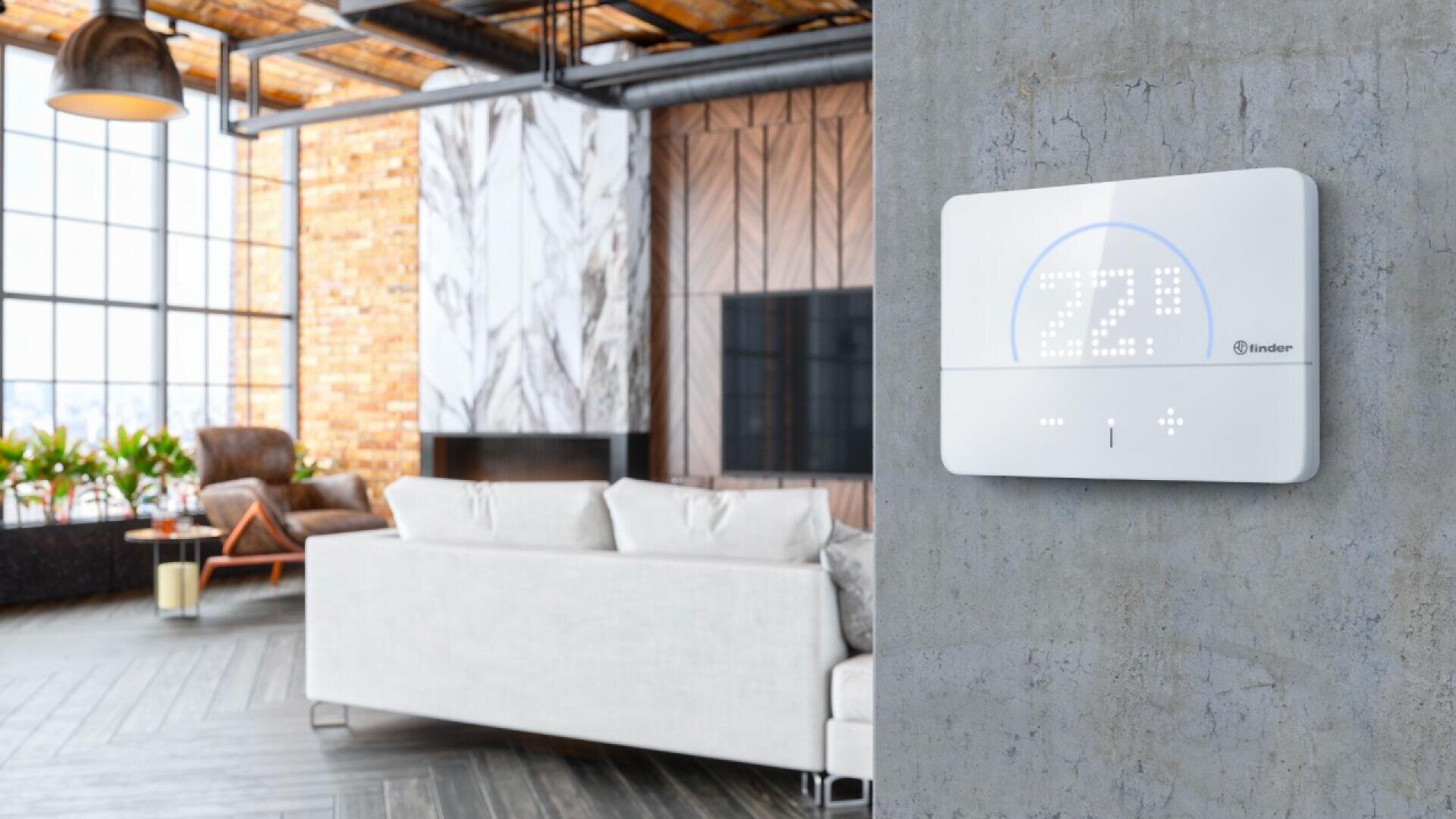 Un thermostat numérique moderne monté sur un mur gris affiche 72°F dans un salon industriel élégant avec un canapé, un fauteuil, des plantes et de grandes fenêtres en arrière-plan.