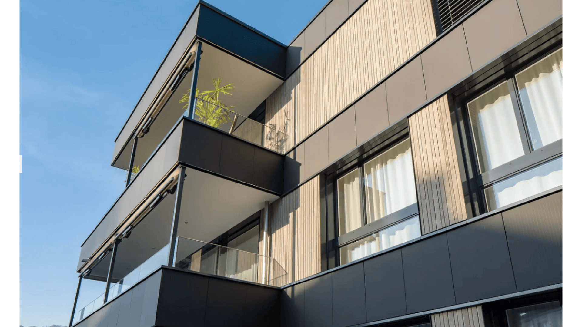 Immeuble d'appartements moderne avec des panneaux en bois et en métal foncé, avec de grandes fenêtres, des balustrades en verre et une plante en pot sur un balcon supérieur, le tout sur fond de ciel bleu clair.