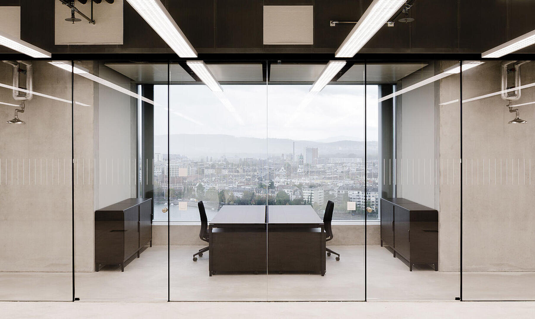 Bureau moderne aux parois vitrées, comprenant deux bureaux noirs, trois chaises et une grande fenêtre donnant sur un paysage urbain où se dessinent, au loin, des immeubles et des collines. La pièce présente un style minimaliste et est bien éclairée.
