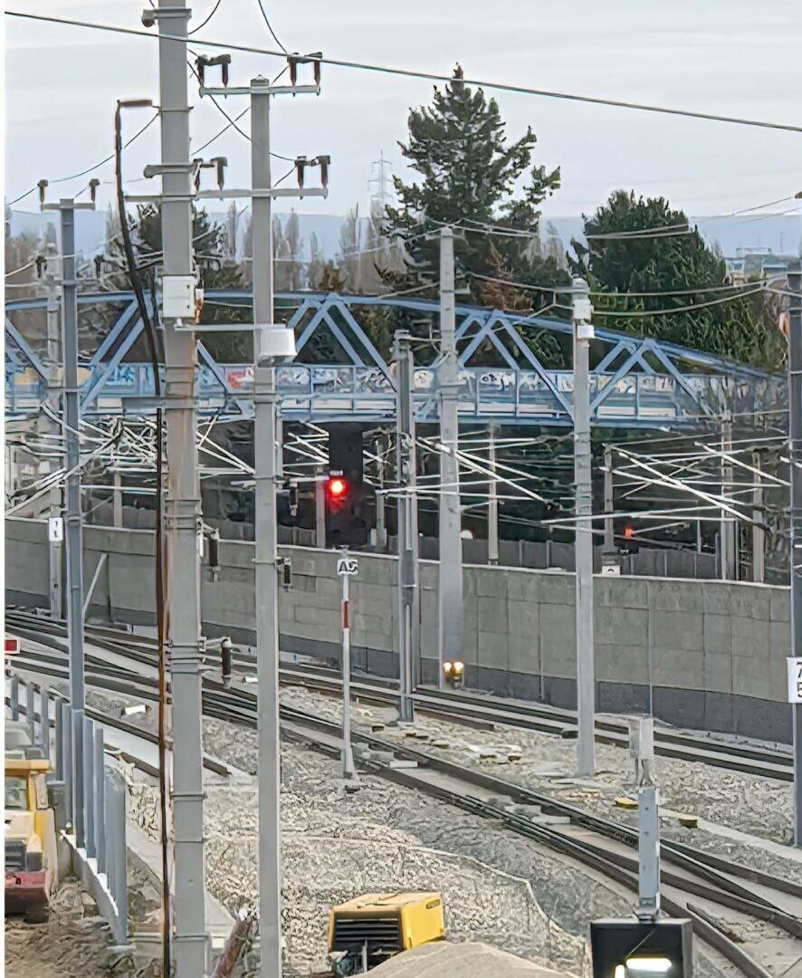 Ein Bahngleis mit Oberleitungen, Masten, einer roten Ampel, Baumaschinen und einer blauen Fußgängerbrücke im Hintergrund an einem bewölkten Tag.