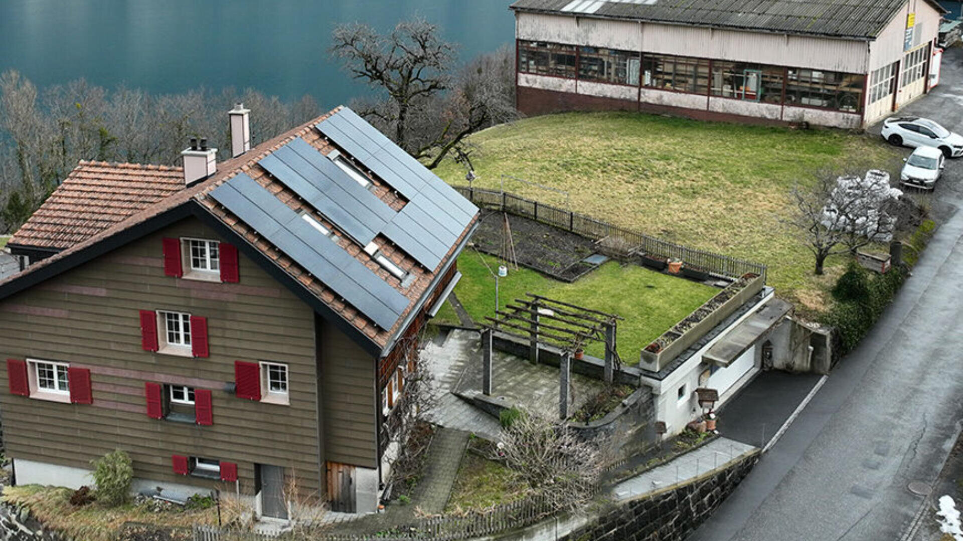 Ein Haus mit Sonnenkollektoren auf dem Dach steht neben einem grünen Hof, einem kleinen Garten und einem größeren Industriegebäude, in der Nähe eines Sees und einer Straße mit geparkten weißen Autos. Bäume und Gras umgeben die Gebäude.