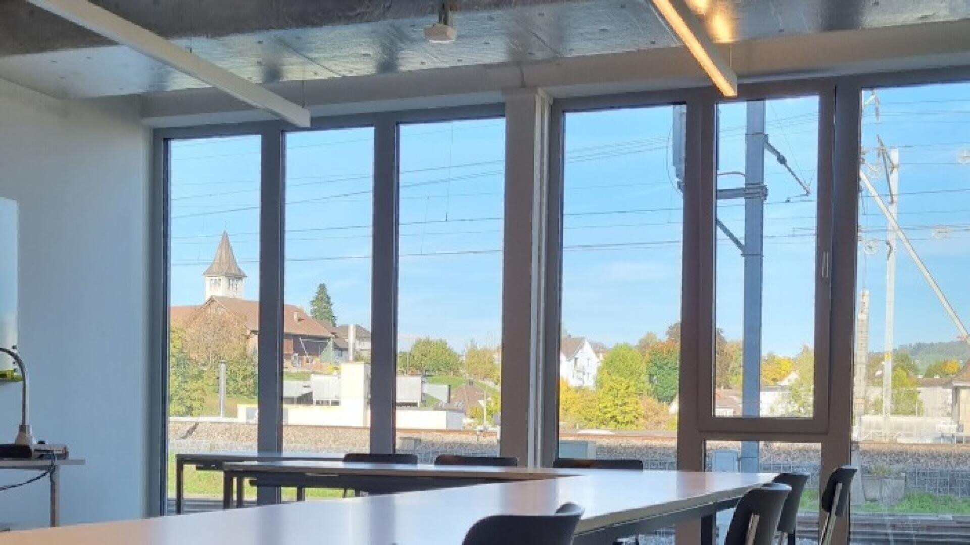 Une salle de classe baignée de soleil, avec des tables et des chaises vides ; de grandes fenêtres donnant sur des arbres, des lignes électriques et des maisons au loin, avec le clocher d’une église sous un ciel bleu.