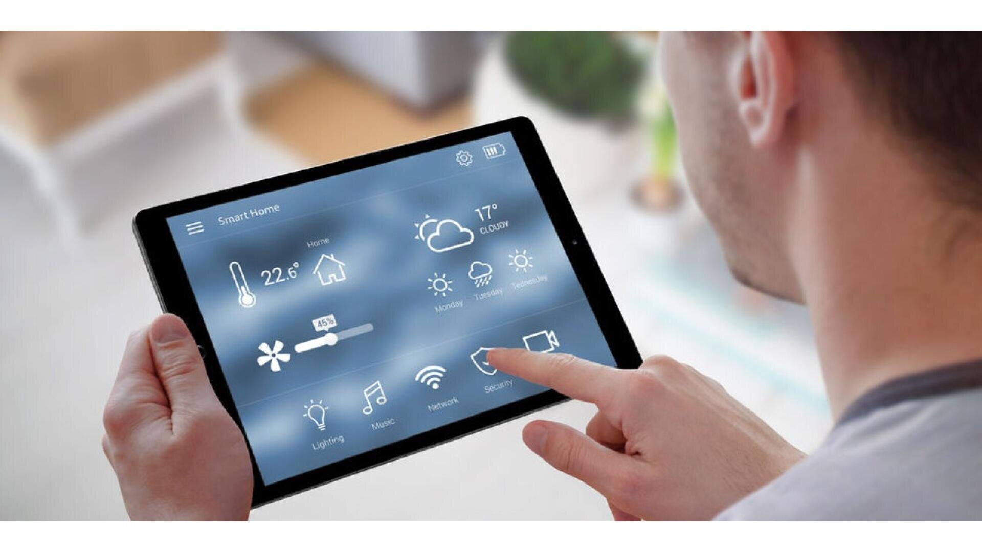 Une personne tient une tablette affichant un tableau de bord de contrôle de maison intelligente, avec des icônes pour la température, l'éclairage, la musique, la météo, le réseau et les paramètres de sécurité. L'écran affiche le temps qu'il fait et la température intérieure.