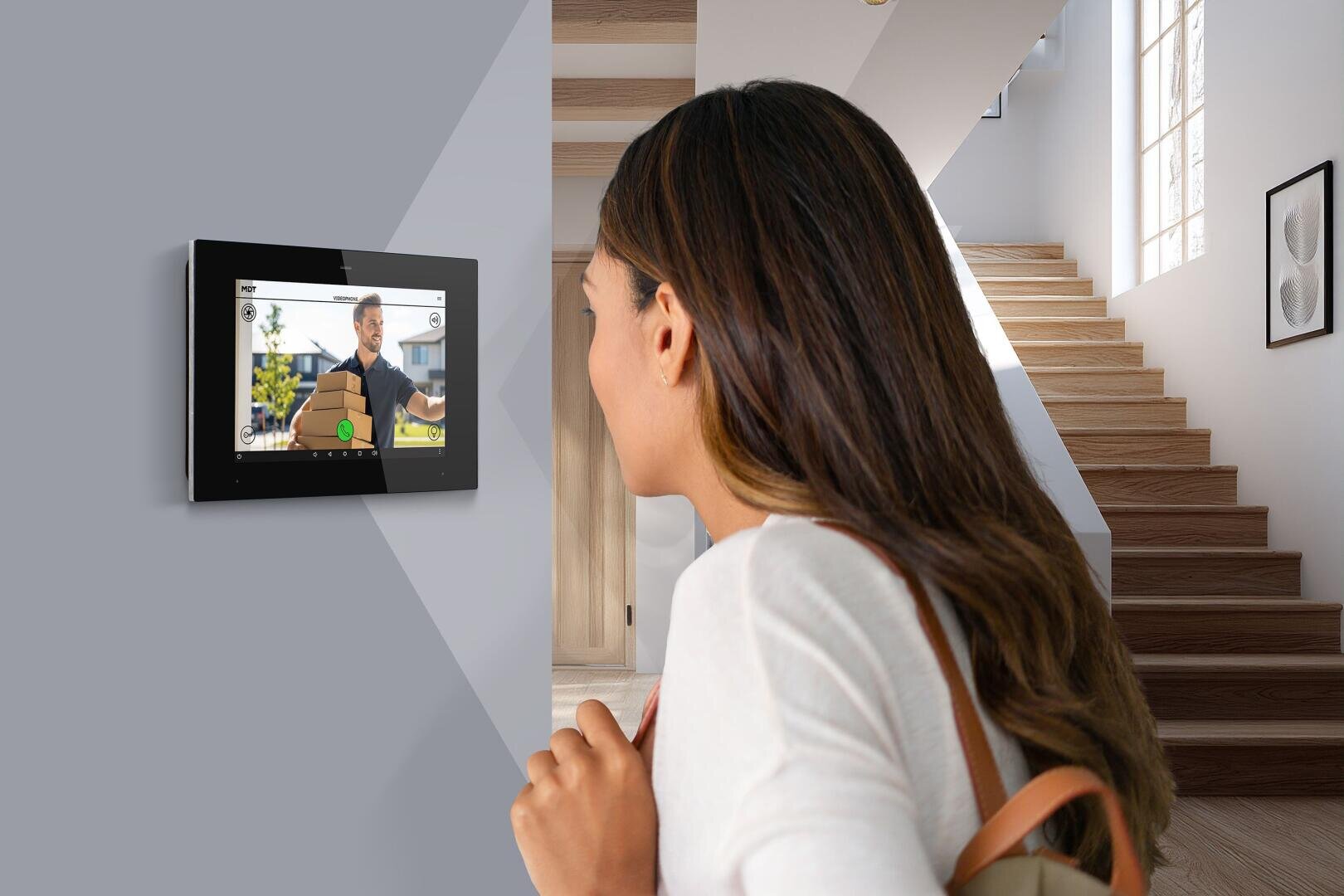 Eine Frau mit langen braunen Haaren steht in einem Haus und blickt auf einen an der Wand befestigten Bildschirm, auf dem ein Mann per Videoanruf spricht. In dem modernen, hellen Hintergrund sind eine Treppe und ein großes Fenster zu sehen.