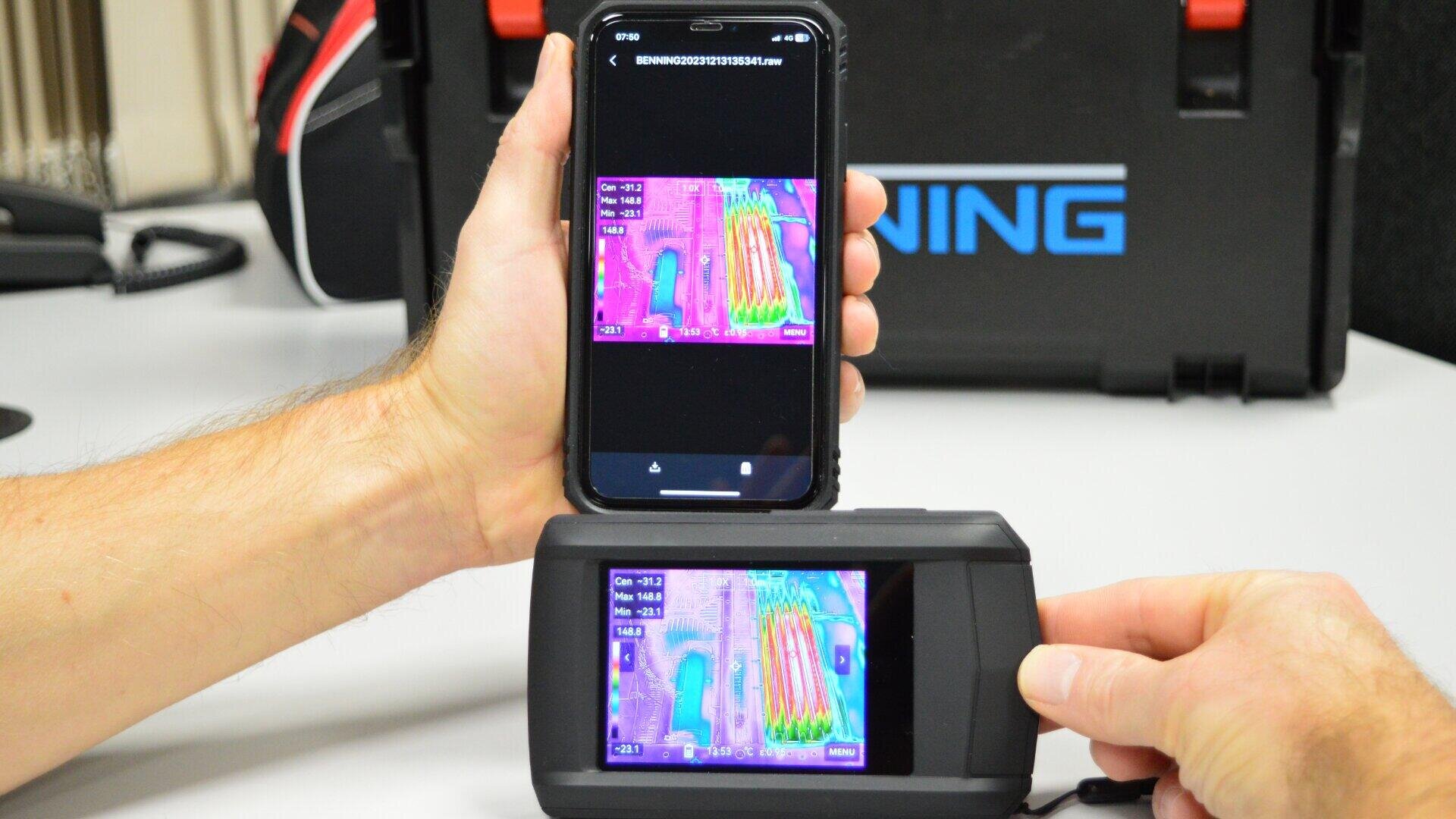 Eine Person hält ein Smartphone in der Hand, auf dem ein Wärmebild angezeigt wird, das identisch mit dem eines tragbaren Wärmebildgeräts ist. Beide Bildschirme zeigen Wärmesignaturen in Rosa-, Lila-, Blau- und Gelbtönen auf einem Schreibtisch.