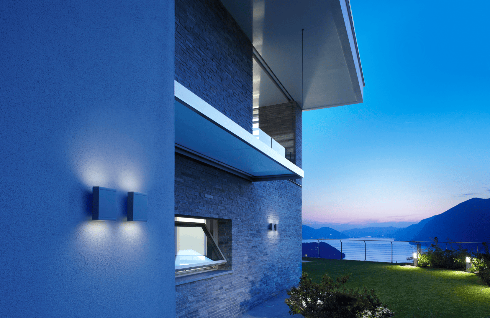 Fassade eines modernen Hauses in der Abenddämmerung mit blauer Beleuchtung, Steinmauern und großen Fenstern; von einem Balkon aus bietet sich ein malerischer Blick auf Berge, Wasser und einen Rasen, im Hintergrund der klare Abendhimmel.