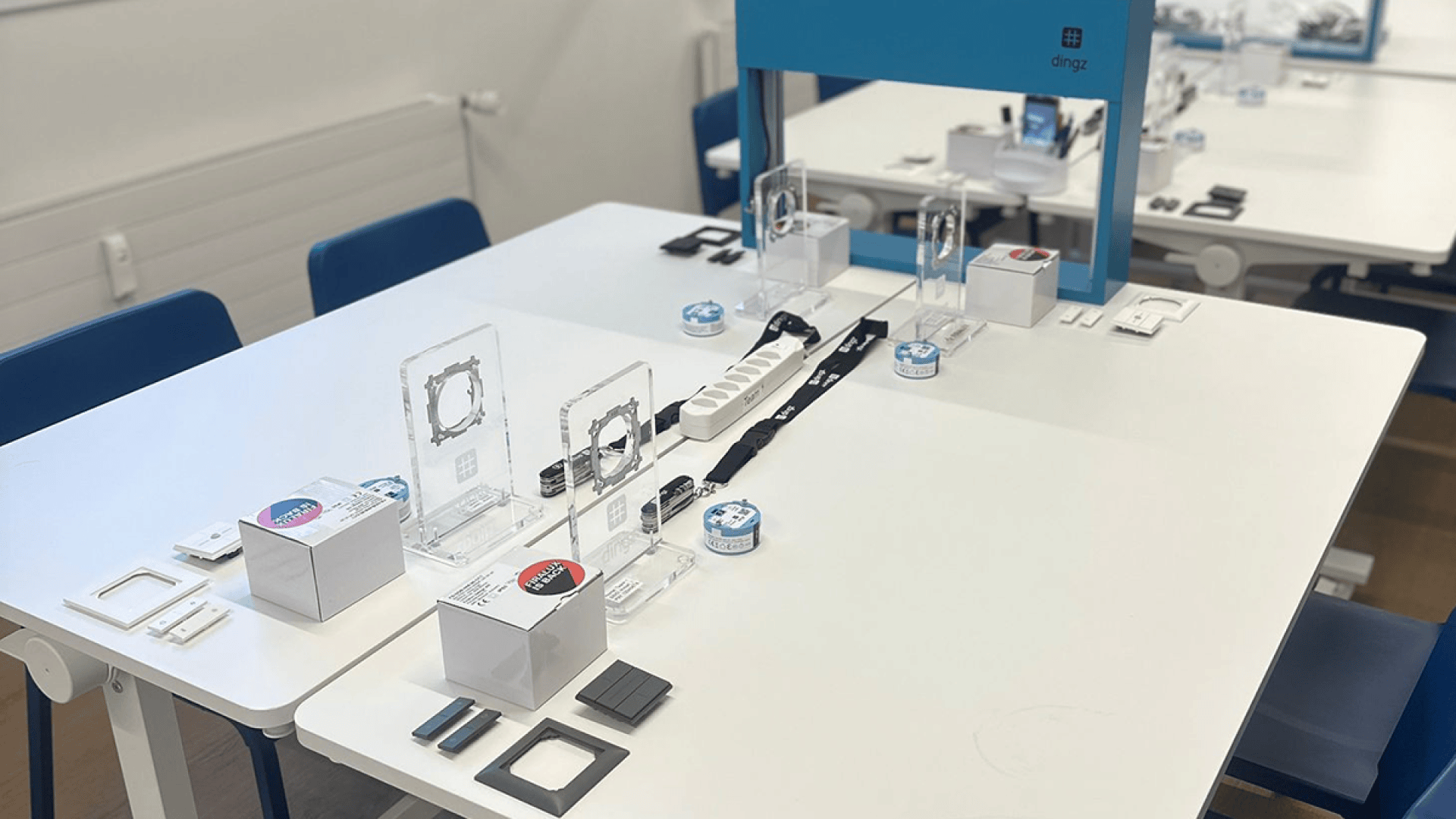 Une salle de classe équipée de tables blanches sur lesquelles sont disposés des kits électroniques, des cadres en plastique transparent, de petits appareils ronds et des chaises bleues, le tout aménagé pour une activité pratique ou un atelier.