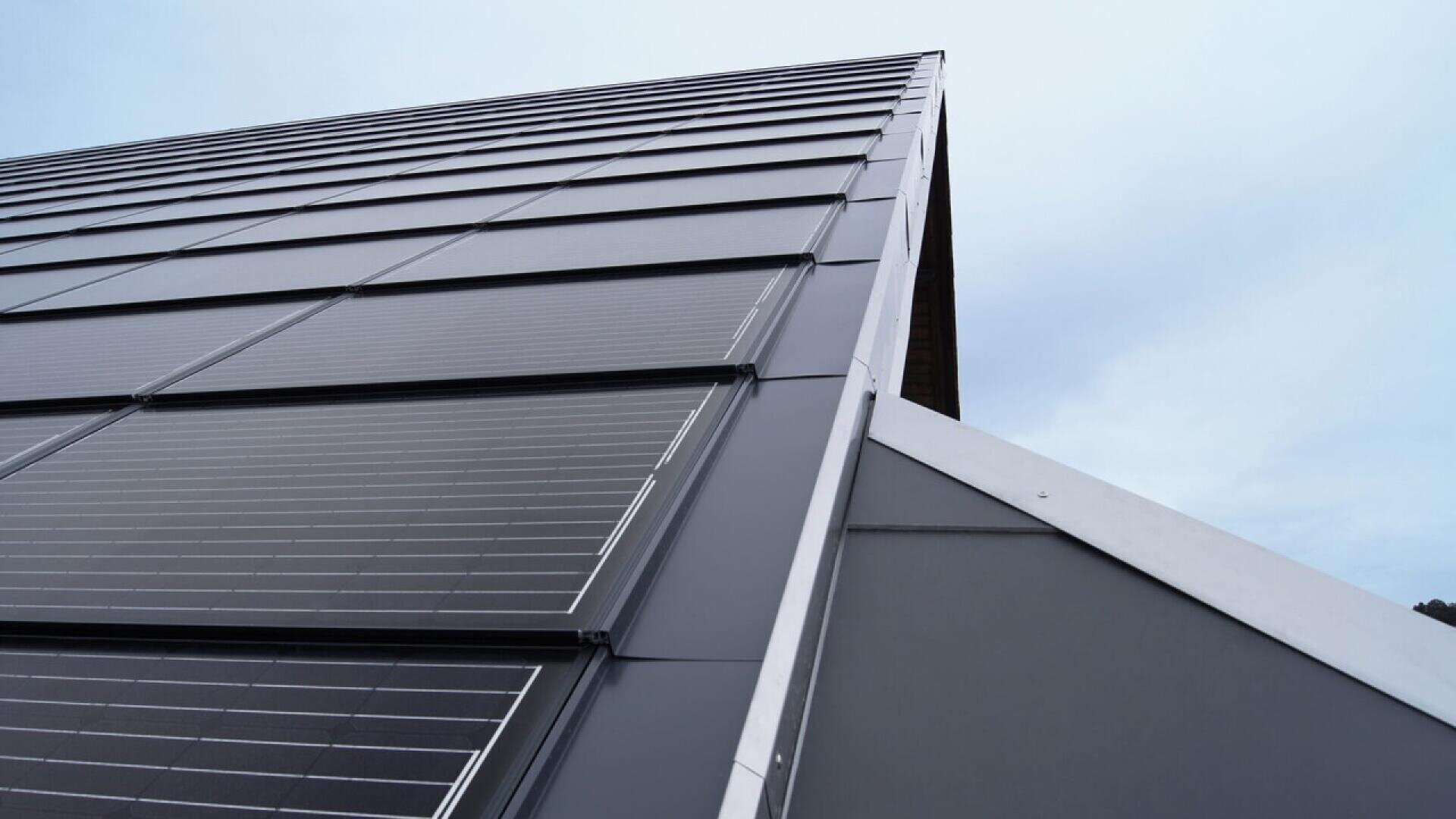 Gros plan sur le toit d'un bâtiment moderne recouvert de panneaux solaires sombres et élégants, sous un ciel nuageux. Les panneaux sont disposés en rangées bien ordonnées le long du toit incliné.