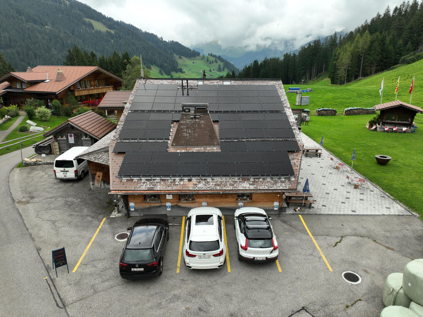 Ein chaletartiges Gebäude mit einer großen Reihe von Solarzellen auf dem Dach, drei davor geparkte Autos und eine grüne Berglandschaft im Hintergrund unter einem bewölkten Himmel.