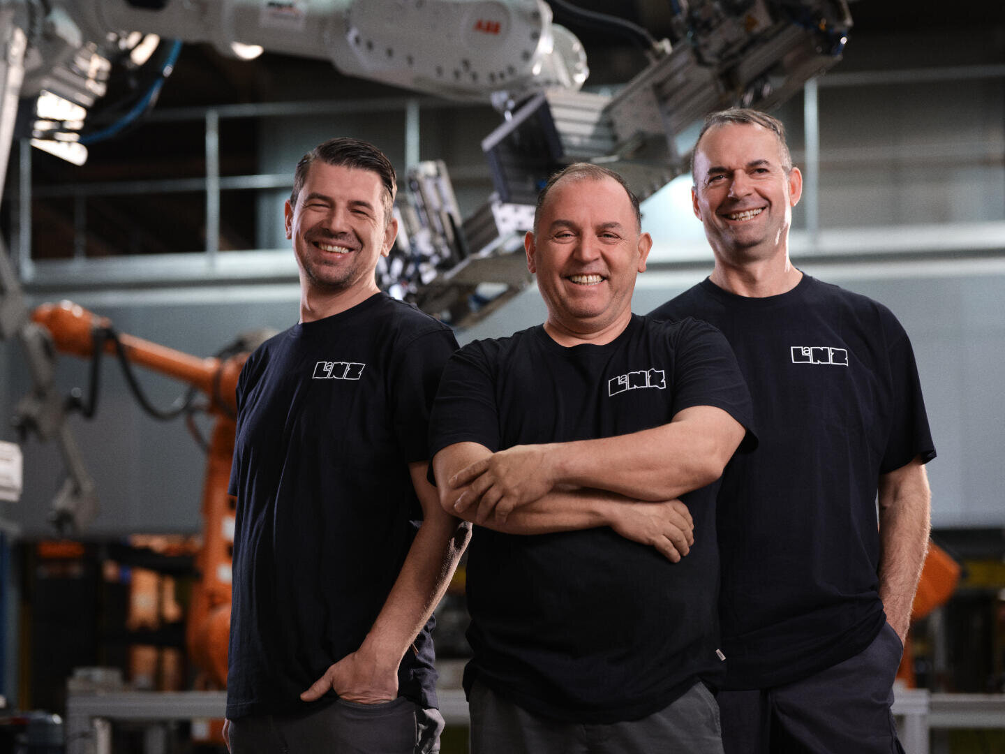 Drei Männer in einheitlichen schwarzen T-Shirts stehen lächelnd vor industriellen Roboterarmen in einer Fabrikumgebung. Die Männer stehen dicht beieinander und wirken freundlich und selbstbewusst.