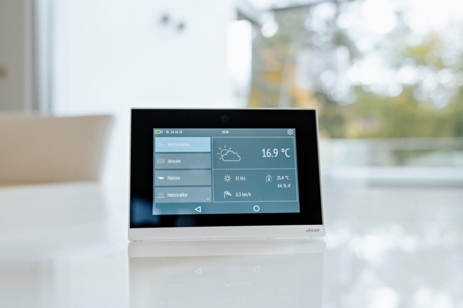 Eine digitale Wetterstation zeigt auf einem Touchscreen eine Temperatur von 16,9 °C, die Wetterbedingungen und andere Informationen an. Das Gerät steht auf einer weißen Fläche in einem hellen, modernen Raum mit großen Fenstern und verschwommenem Grün draußen.