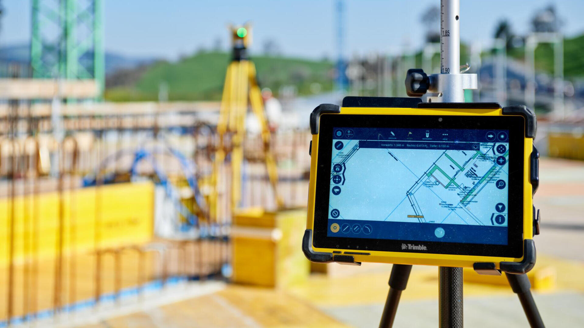 Un appareil d'arpentage numérique avec un écran affichant des plans de construction est monté sur un trépied sur un chantier de construction, avec du matériel de construction et des structures floues à l'arrière-plan.