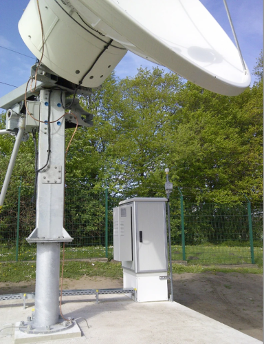 Une grande antenne parabolique montée sur un support métallique se trouve à l'extérieur, près d'un terrain clôturé avec des arbres verts et une armoire à outils grise en dessous. Le ciel est clair et bleu.