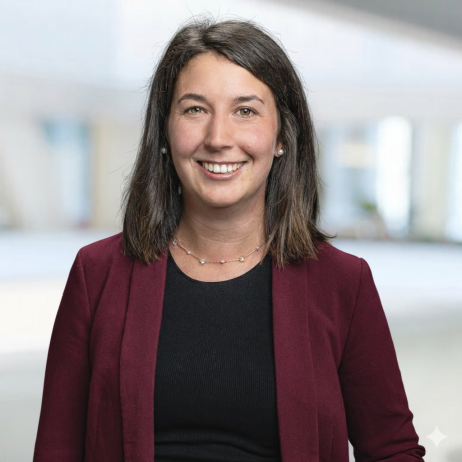 Eine Frau mit glattem braunem Haar und einem freundlichen Lächeln trägt einen weinroten Blazer über einem schwarzen Oberteil. Sie steht in einem hellen, verschwommenen Innenraum, möglicherweise einem Büro.