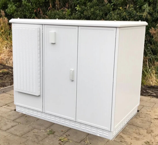 Une grande armoire métallique blanche à trois portes est placée à l'extérieur sur une surface pavée, avec de la verdure et des plantes en arrière-plan.