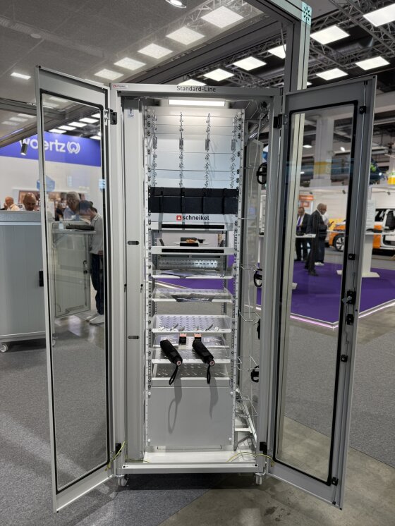 Ein hohes Serverrack aus Glas mit offenen Türen, in dem elektronische Geräte und Kabel untergebracht sind, ausgestellt auf einer Technikmesse. Im Hintergrund sind Menschen und Messestände zu sehen.