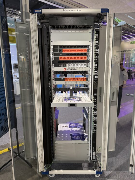 Server-Rack mit beschrifteten Schaltern, Sicherungsautomaten und elektrischen Bauteilen, ausgestellt an einem Messestand, dessen Glastüren geöffnet sind, um die Verkabelung und den Aufbau im Inneren zu zeigen.