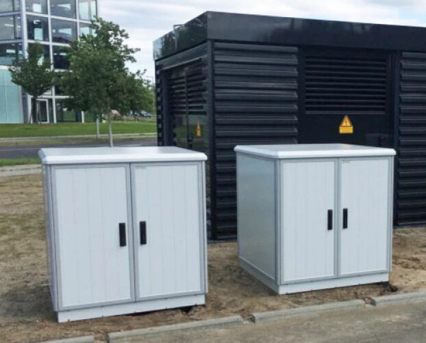 Deux boîtiers électriques gris clair à double porte sont placés sur un socle en béton à l'extérieur, devant un bâtiment industriel noir plus grand. En arrière-plan, on aperçoit des arbres et un bâtiment moderne en verre.