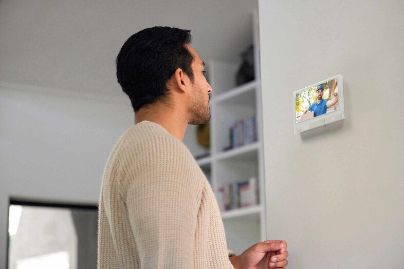 Un homme se tient à l'intérieur et regarde un écran d'affichage intelligent mural montrant un appel vidéo avec deux personnes qui se sourient et se font des signes de la main. Des étagères contenant des livres et des objets de décoration sont visibles à l'arrière-plan.