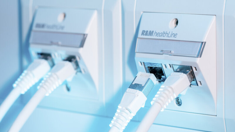Zwei weiße Ethernet-Kabel sind an Wand-Netzwerkanschlüsse mit der Aufschrift „R&M healthLine“ angeschlossen, und eine blaue Beleuchtung hebt diese Geräte hervor.