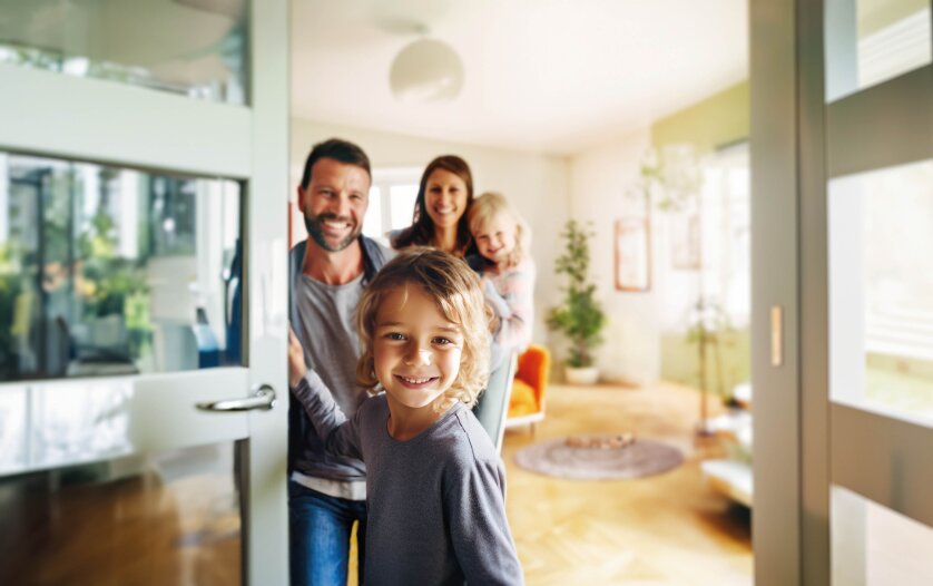 Une jeune fille souriante ouvre une porte vitrée, accueillant le spectateur à l'intérieur. Derrière elle, deux adultes et un enfant se tiennent ensemble dans un salon moderne et lumineux, agrémenté de plantes et de lumière naturelle.