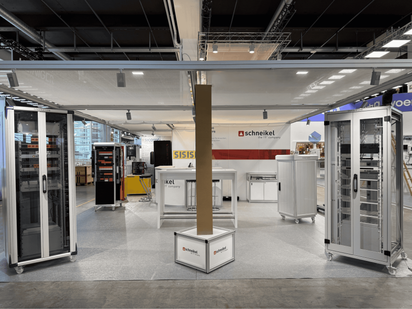 Messestand mit Server-Racks, elektronischen Geräten und Schränken. Im Hintergrund des Standes ist ein rot-weißes Schneikel-Banner zu sehen, und um eine zentrale Säule herum sind verschiedene Gehäuse angeordnet.