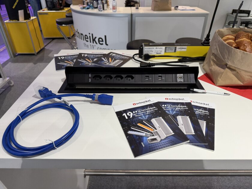 Auf einem weißen Tisch an einem Messestand liegen eine Steckdosenleiste, ein blaues Kabel, Broschüren und ein Stift. Auf der rechten Seite des Bildes ist außerdem eine Papiertüte mit Gebäck zu sehen.