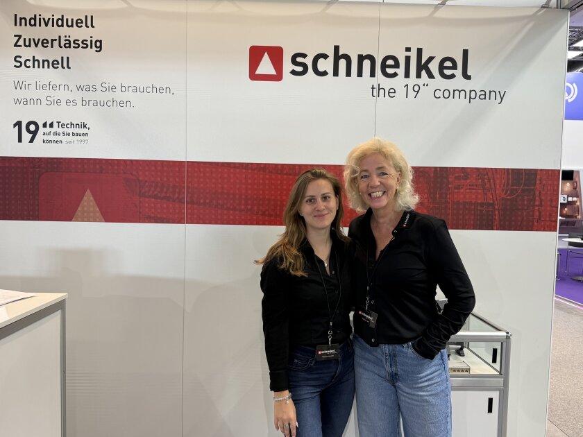 Zwei Frauen stehen an einem Messestand von „schneikel, the 19 company“ und lächeln in die Kamera; im Hintergrund ist eine Wand mit deutschem Text zu sehen, und neben ihnen sind Geräte ausgestellt.