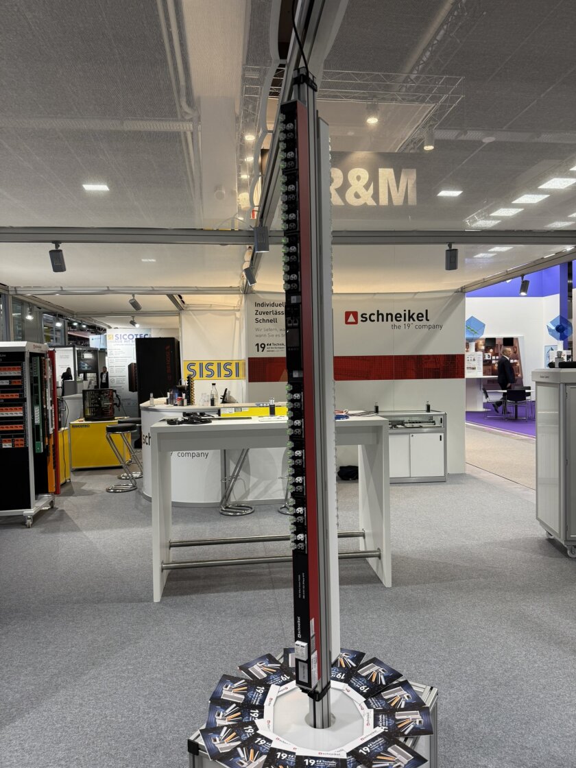 Ein hohes, schlankes elektronisches Display mit Schaltern wird an einem Messestand präsentiert, im Hintergrund sind Schilder mit den Aufschriften „Schneikel“ und „R&M“ zu sehen; daneben liegen Broschüren auf dem Bodenständer.