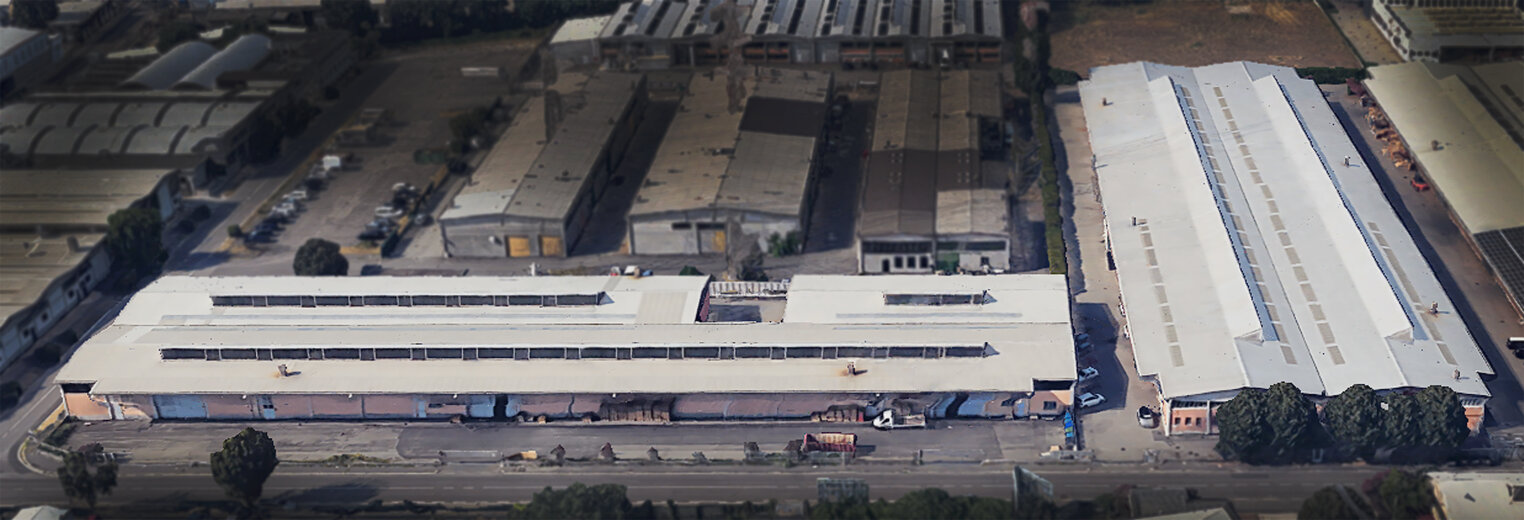 Luftaufnahme von großen Industrielagern mit weißen Dächern, umgeben von Bäumen und einer Straße, in einem städtischen Industriegebiet mit weiteren Fabrikgebäuden in der Nähe.