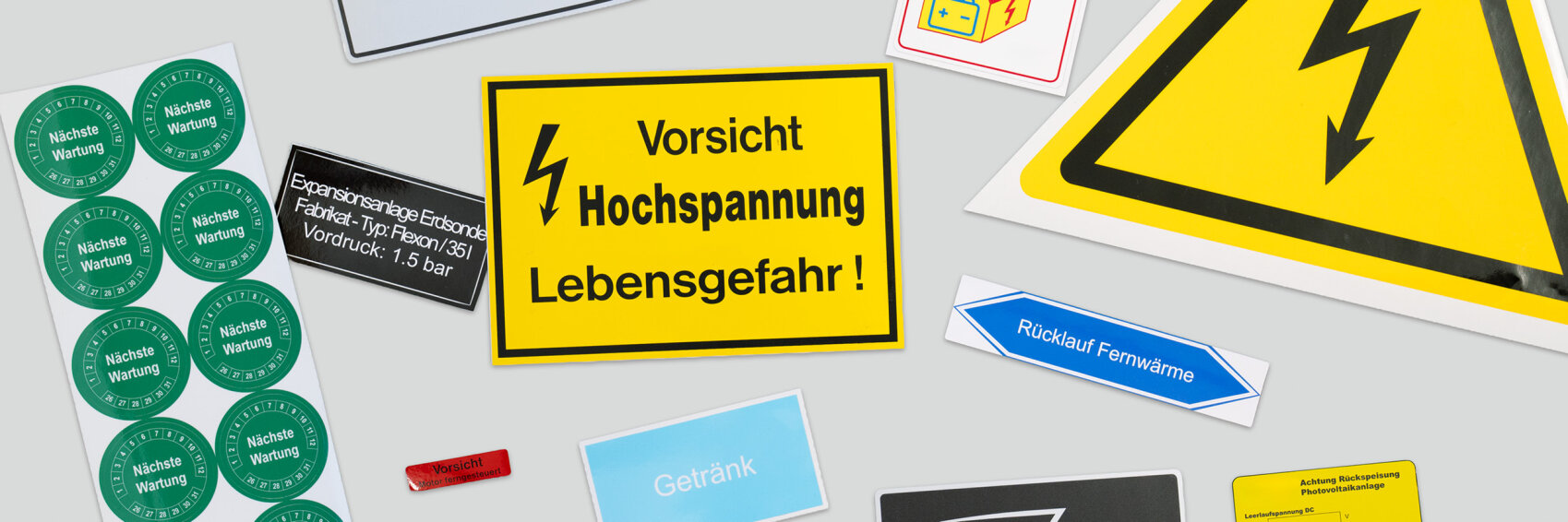 Sur un fond clair, divers panneaux d'avertissement et d'information sont visibles, dont un grand panneau jaune portant l'inscription « Vorsicht Hochspannung Lebensgefahr ! » et un symbole d'éclair.