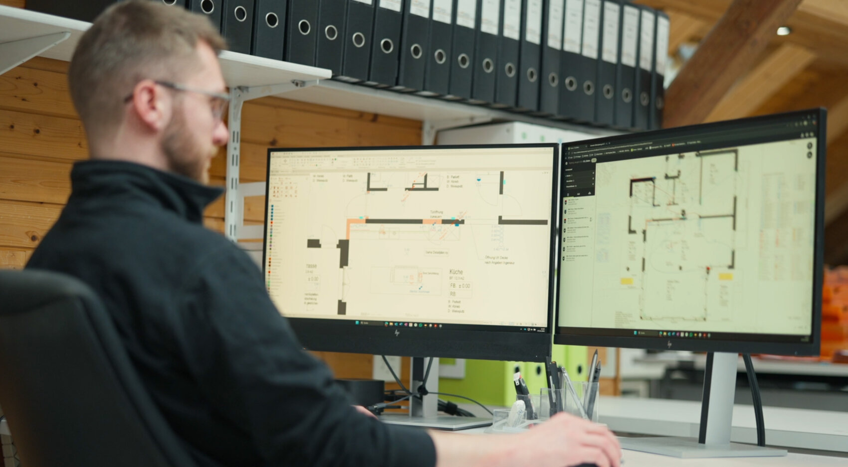 Eine Person sitzt an einem Schreibtisch und arbeitet an zwei Computermonitoren, auf denen architektonische oder technische Grundrisse angezeigt werden, während im Hintergrund Regale mit schwarzen Ordnern zu sehen sind.
