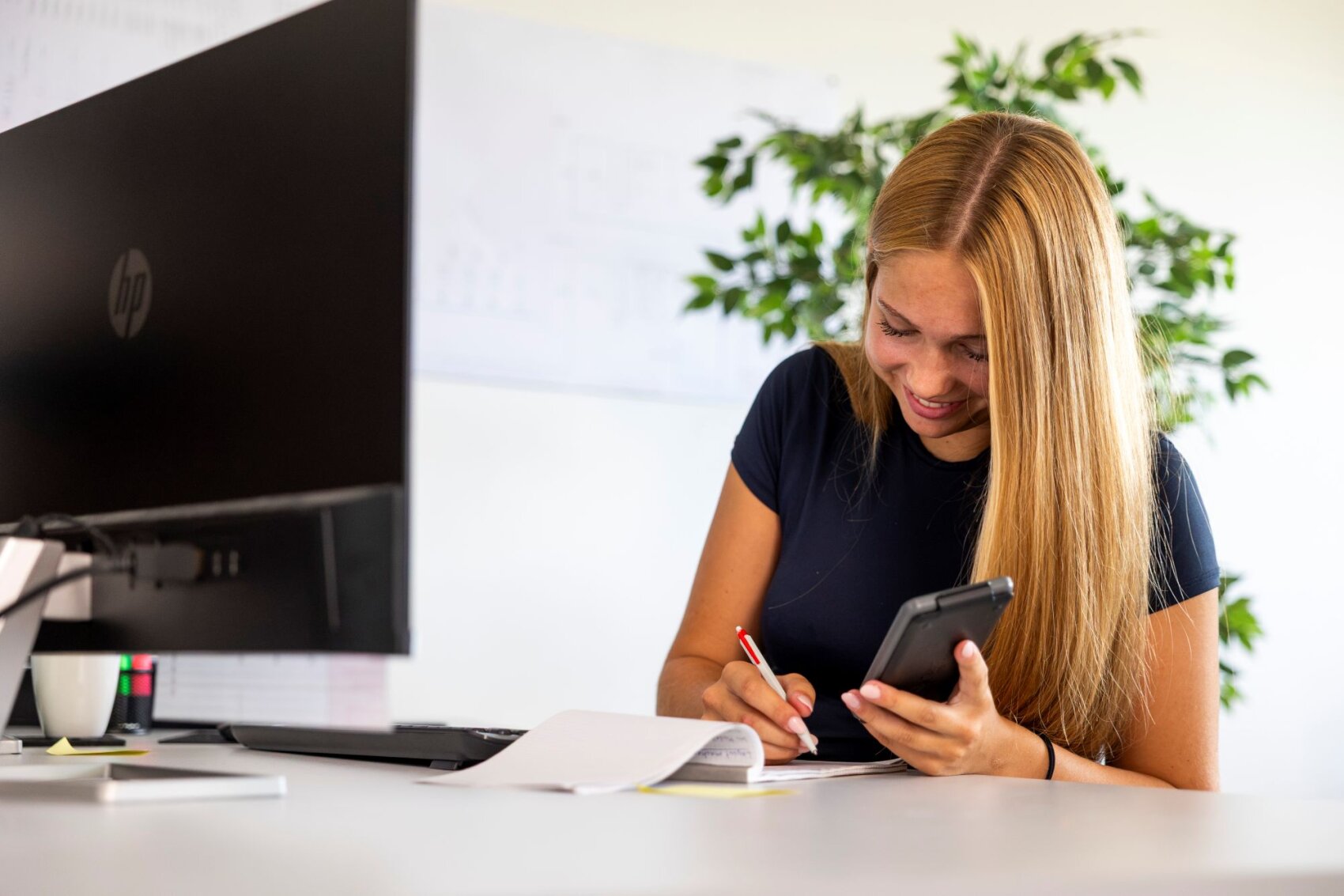 Une femme aux longs cheveux blonds sourit tandis qu'elle travaille à son bureau, une calculatrice à la main, en écrivant sur une feuille de papier ; à l'arrière-plan, on aperçoit un écran d'ordinateur, des documents et une plante.
