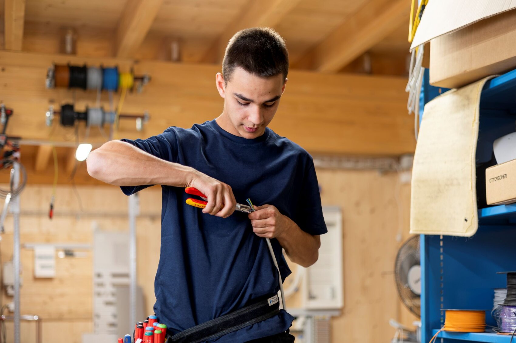 Un jeune homme vêtu d'une chemise bleu marine travaille sur un câble à l'aide d'une pince dans un atelier aux murs en bois, où sont rangés des outils et des caisses sur des étagères.